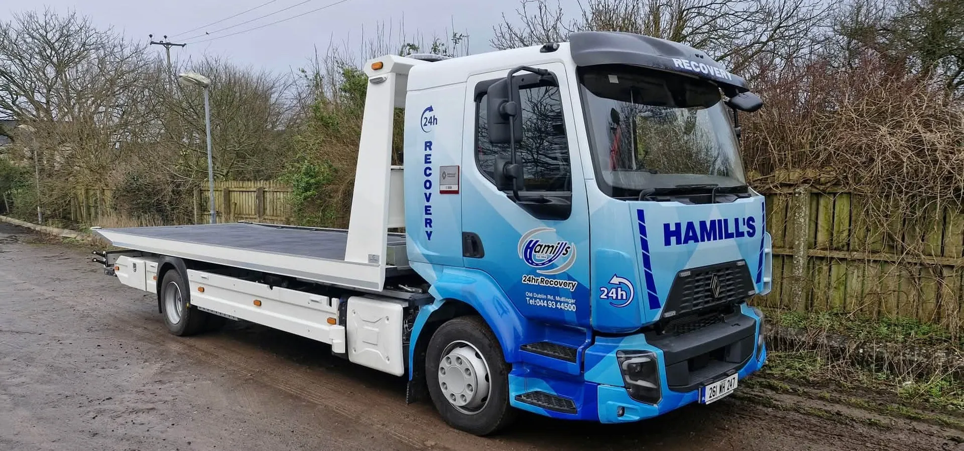 Hamill Group flatbed recovery truck in a blue geometric vinyl wrap, parked on a rural road.